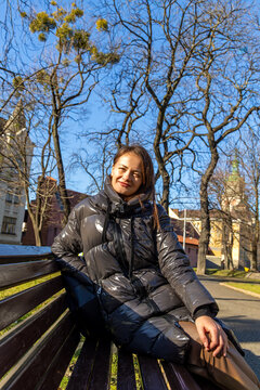 Smiling Woman Sitting on Park Bench in Winter Sunlight. Smiling woman in a shiny black puffer jacket sits on a wooden park bench in warm sunlight beneath leafless trees in an urban park.