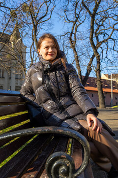 Smiling Woman Relaxing on Park Bench in Sunny Weather. Smiling woman in a shiny black puffer jacket sits on a wooden park bench in bright sunlight with leafless trees and city buildings behind.
