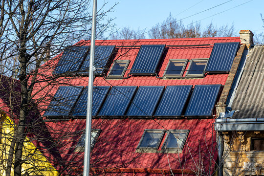 Solar Collectors Installed on Red Roof with Skylights. Solar thermal collectors and skylights cover a red metal roof beneath a clear blue sky with bare tree branches in front.