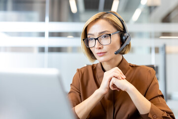 Confident businesswoman wearing a headset and glasses, working on a laptop in an office, actively...