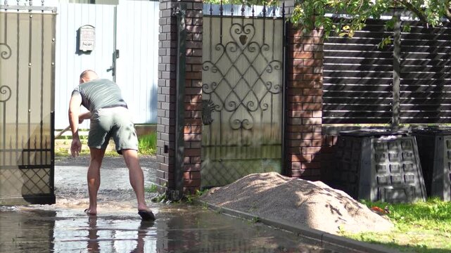 Man uses a large squeegee broom to push muddy flood water away from his house entrance after heavy rain.