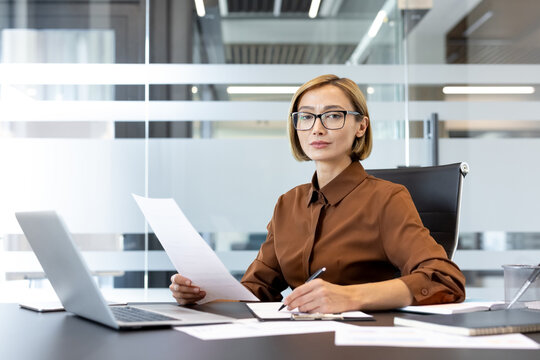 Confident executive woman working at her desk, holding documents and writing notes, managing paperwork and responsibilities in a contemporary office environment