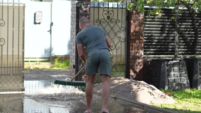 Man uses a large squeegee broom to push muddy flood water away from his house entrance after heavy rain.