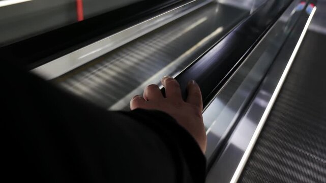 Close-up view of a person's hand holding a black rubber handrail while riding a moving escalator travelator. Metal steps with LED lighting visible in a modern station, airport, or mall interior.
