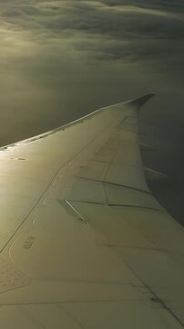 Airplane wing banking turning over thick dramatic clouds. Flaps changing angle during aircraft maneuver, sunlight shifting fade out on wing surface. Passenger plane window view banking left.