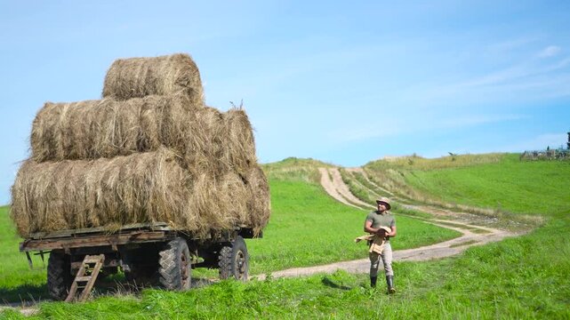 Highly engaging commercial ready wide shot of a confident male farmer in a bucket hat walking forward next to a large loaded hay wagon under a bright summer sky.