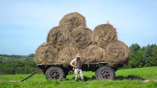Highly engaging commercial ready wide shot of a satisfied male farmer patting and checking fresh round hay bales stacked on a rustic trailer.