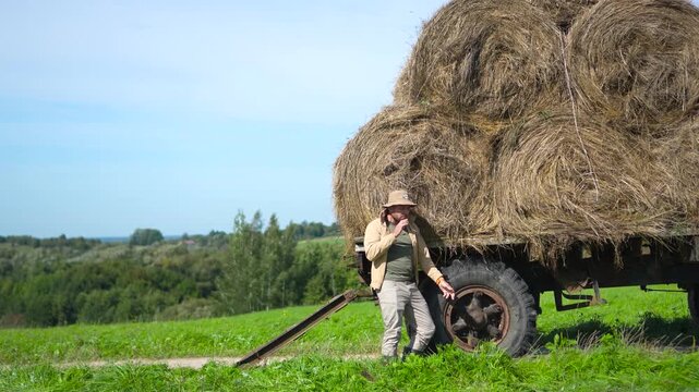 Highly engaging commercial ready medium shot of a male farmer standing by a rustic hay wagon, picking a straw and putting it in his mouth while relaxing in a green field.