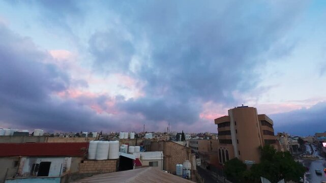 Morning view over Madaba, Jordan, with dense low-rise buildings and rooftop water tanks beneath dramatic blue and pink clouds at dawn.