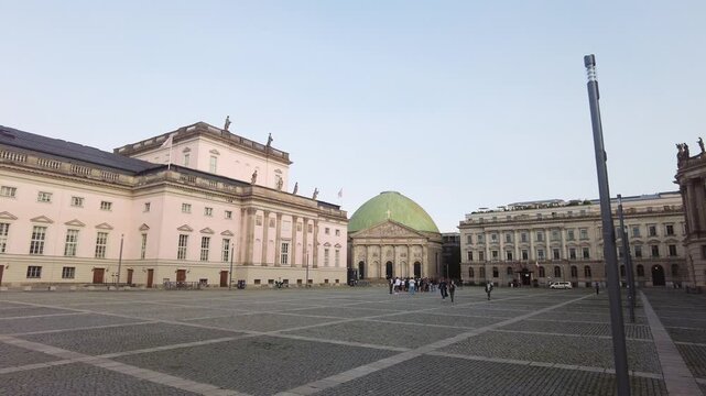 Camera pans right across a cobblestone plaza revealing St. Hedwig's Cathedral dome and State Opera building while pedestrians walk in the distance.
