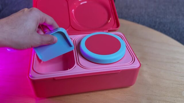 A close-up view of a hand assembling a bright pink bento lunchbox on a wooden table. The person inserts a blue divider into the empty food container indoors under warm creative lighting.