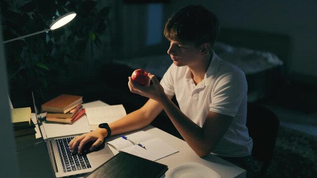 Concentrating adolescent boy eating a fresh red apple at the dark night desk.