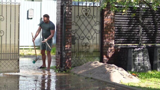 Homeowner holds a broken squeegee broom in his hands while cleaning muddy floodwater from his driveway.