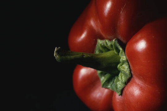 Macro shot of ripe red bell pepper with vibrant green stem in moody lighting on dark background. Fresh raw vegetable for organic recipes, culinary menus, nutrition, and healthy eating.