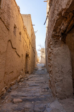 Empty narrow alley winding between weathered mudbrick houses toward distant buildings, Ad Dakhiliyah Region, Al Hamra, Oman