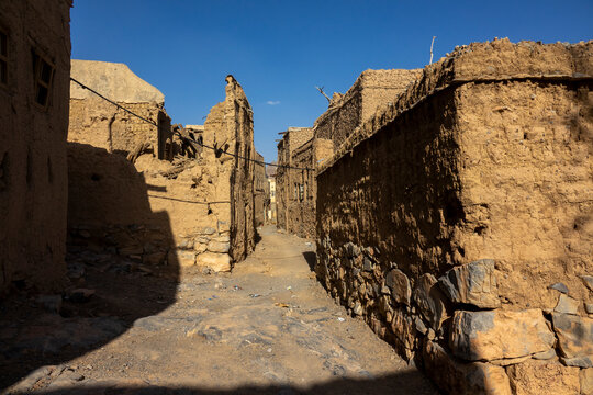 Narrow alley lined with ruined mudbrick walls leading toward distant buildings, Ad Dakhiliyah Region, Al Hamra, Oman