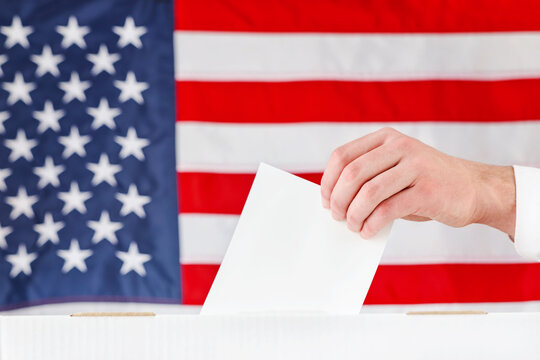Hand holds a blank ballot over a voting box before an American flag
