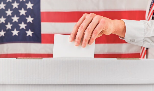 Hand inserts a blank ballot into a box in front of an American flag