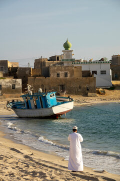 Man in a white dishdasha standing on the beach and looking toward a beached fishing boat and village with a green mosque minaret, Dhofar Governorate, Mirbat, Oman