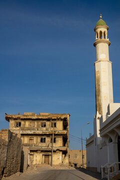 Ruined multi-story building standing beside a white mosque minaret with a green dome under a clear blue sky, Dhofar Governorate, Mirbat, Oman