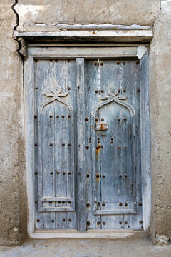 Old weathered wooden double door with a carved arch motif metal studs and a hanging padlock set in a mudbrick wall, Dhofar Governorate, Mirbat, Oman