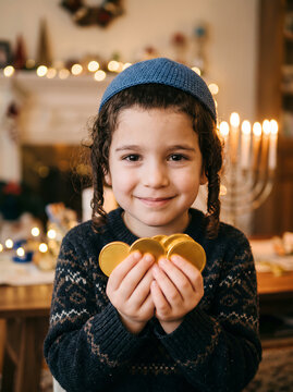 Smiling Jewish boy wearing blue kippah holding gold chocolate gelt coins with menorah in background. Hanukkah or Chanukah celebration concept with copy space.
