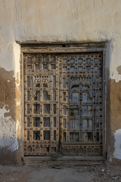Ornately carved wooden double door with geometric metal studs and an Arabic inscription set in a weathered plaster wall, Dhofar Governorate, Mirbat, Oman