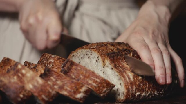 Close-up of baker in rustic linen apron slicing freshly baked artisan sourdough bread with knife on wooden cutting board. Homemade crusty loaf for breakfast, bakery promos, and healthy food concepts.