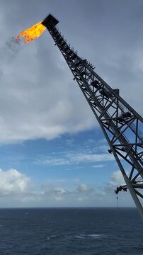 Industrial gas flare stack on an offshore oil rig burning natural gas against a cloudy blue sky over the sea.