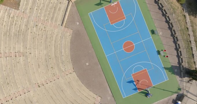Aerial zenithal view of an empty outdoor basketball court with a blue playing surface, next to the curved concrete steps of a small grandstand, seen from above on a sunny day. The park is empty.