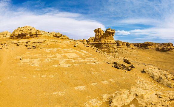 An aerial view of yellow yardang rock formations under bright sunlight with soft clouds across a blue sky in Xinjiang, China.