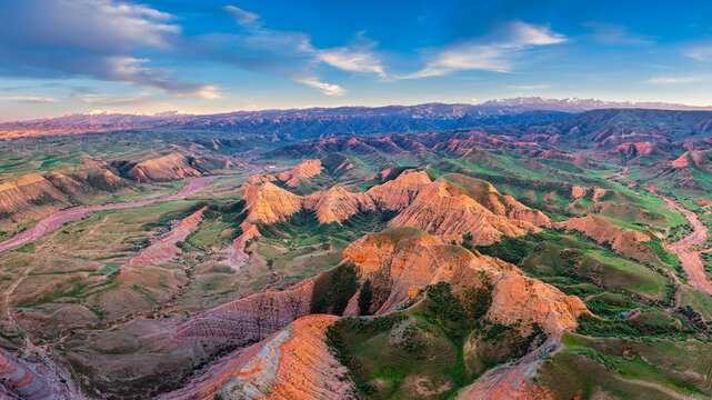 Aerial view of colorful Danxia landform mountains with green valley vegetation and distant snow peaks under bright sunlight.