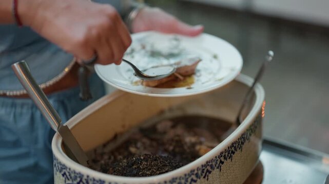 Hand serving hot food from large pot into plate at buffet counter with ladle