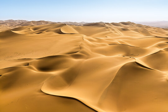 Vast golden sand dunes stretching across the desert landscape under bright sunlight and a clear blue sky in Xinjiang, China.