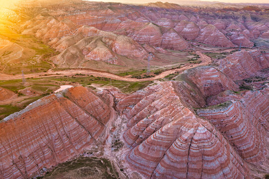Aerial view of colorful red danxia landform mountains with stratified rock layers and winding riverbeds under warm golden sunlight in Xinjiang, China.