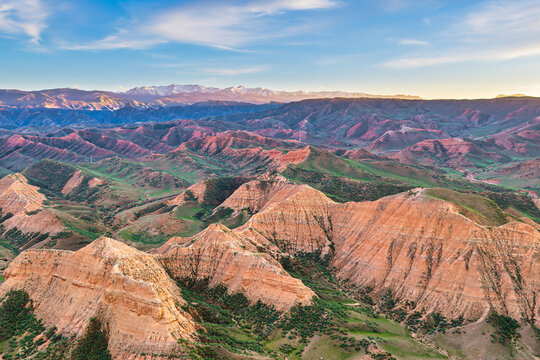 Aerial view of layered reddish rock formations and green slopes under bright sunlight with distant snow-capped mountains and a blue sky in Xinjiang.