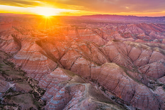 Aerial view of red and orange layered landforms under a bright sunset sky in Xinjiang, China, with deep valleys and ridges bathed in warm sunlight.