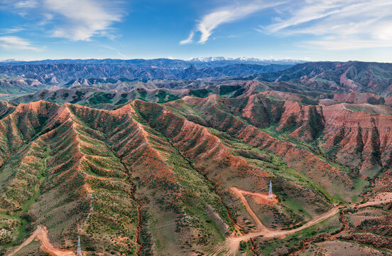 High-angle landscape shows rugged red mountain ridges covered in sparse vegetation under a blue sky with thin clouds in bright daylight.