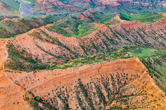 Aerial view of red rock mountains with steep cliffs, layered ridges, and green vegetation illuminated by bright daylight in Xinjiang, China.