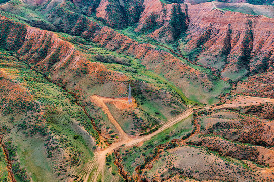 High angle view of rugged red and green landforms in Xinjiang, displaying layered rock erosion, dirt tracks, and scattered vegetation under soft daylight.
