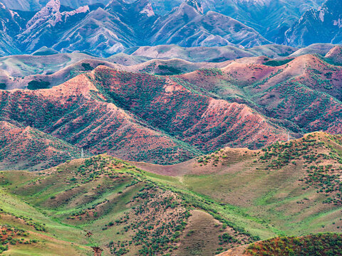 Layered mountain range displays reddish soil slopes, patchy green vegetation, and blue distant peaks under soft daylight in Xinjiang.