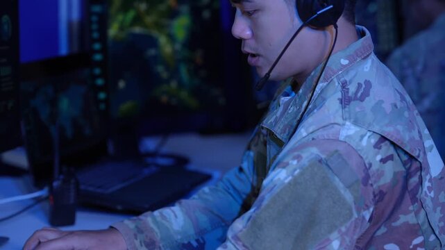 Focused military operator in camouflage uniform wearing a headset and using a computer in a dim blue-lit command center with server racks.