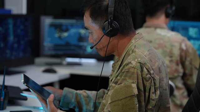 Military operator wearing a headset checks a tablet in a dark command center, with another uniformed colleague and digital maps on screens behind him.