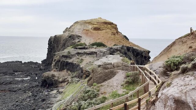 A girl walks on a wooden footpath, admiring the rugged cliff landscape and waves crashing against the rocks at Cape Schanck, Melbourne, Mornington Peninsula, Victoria, Australia.	