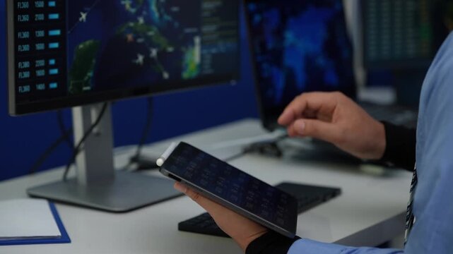 Person holding a tablet while monitoring maps and data on multiple screens in a modern control room office.