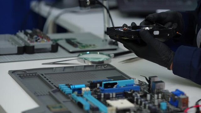 Technician wearing black gloves repairs a hard drive above a computer motherboard on an electronics workbench.