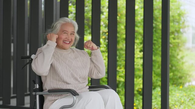 Elderly woman smiles, flexing arms in wheelchair. Background shows green trees behind dark slats. Her joy shows strength and resilience