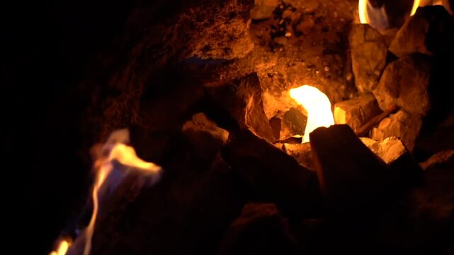Close up shot of bright orange flames and fire tongues burning between dark stones in dark environment.