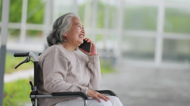 Elderly woman smiles while talking on phone. She sits in wheelchair under bright windows. Her expression shows warmth and joy