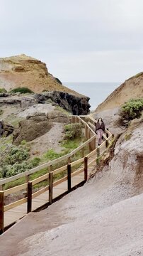 A girl walks on a wooden footpath, admiring the rugged cliff landscape and waves crashing against the rocks at Cape Schanck, Melbourne, Mornington Peninsula, Victoria, Australia.	